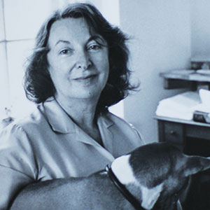 A woman with wavy hair wearing a light-colored blouse smiles at the camera while holding a dog indoors near a desk with papers. The image is in black and white.