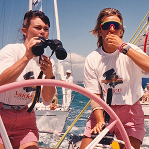 Two women in matching white shirts and pink shorts on a sailboat, one looking through binoculars and the other steering with a pink wheel. Another boat and a sailor are visible in the background.
