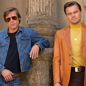 Two men stand side by side in front of a stone pillar. One wears a denim jacket and sunglasses, while the other wears a brown leather jacket over a yellow turtleneck. Both look toward the camera with relaxed expressions.