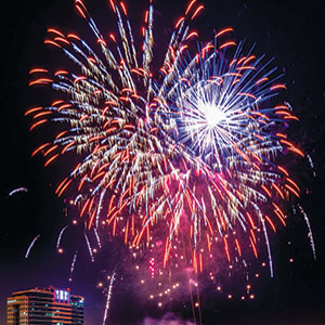 Bright, colorful fireworks burst in the night sky above a lit-up building, creating a vibrant display of red, white, and blue streaks and sparks.