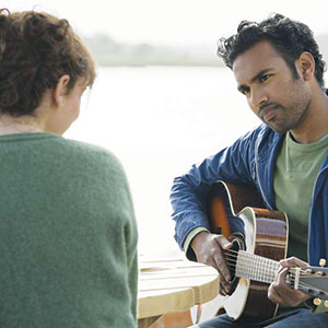 A man sits outdoors playing an acoustic guitar while looking at a woman seated across from him at a wooden table. The background is bright and out of focus, suggesting they are near water or in a park.