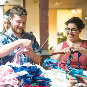Two people smile and work with colorful fabric scraps at a table, appearing to create something together in a bright, indoor setting.