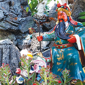 Colorful statue of a fierce, bearded warrior in ornate robes holding a long weapon, surrounded by rocks, greenery, and small plants in an outdoor setting.