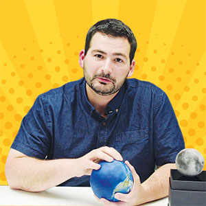 A man with short dark hair and a beard sits at a table holding a model of the Earth; a model of the Moon is on the table beside him. The background is bright yellow with radiating lines and dots.