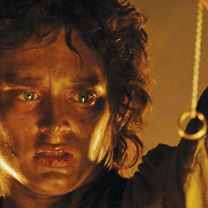 A person with curly brown hair and a dirty face looks intensely at a gold ring hanging on a chain, illuminated by warm light.