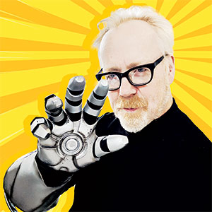 A man with light hair and glasses wears a robotic glove and holds his hand up toward the camera, with a bright yellow sunburst background behind him.