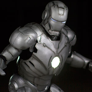 A metallic, armored figure resembling Iron Man stands with glowing eyes and a bright circular light in the chest, set against a dark background.