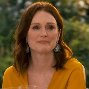 A woman with shoulder-length brown hair, wearing colorful earrings and a yellow top, sits outdoors with greenery in the blurred background. She has a neutral expression on her face.