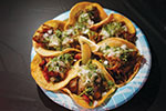A plate with six tacos, each topped with diced onions, cilantro, and a slice of lime in the center. The tacos are arranged in a circle on a blue patterned plate.