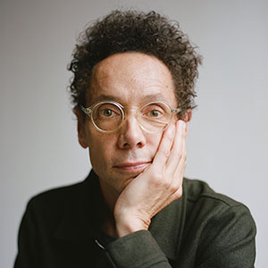 A person with short curly hair, glasses, and a thoughtful expression rests their face on their hand, looking directly at the camera. They are wearing a dark-colored top and sitting against a plain background.