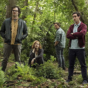Four people stand and sit in a forest clearing surrounded by green foliage; three men are standing, one woman is crouching, and they all have serious expressions.