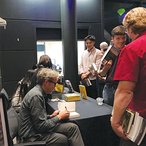 A man sits at a table signing books for a line of people at a book event. Several people in line hold books and smile as they wait their turn. The setting appears to be indoors with a dark background.