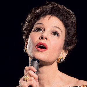 A woman with short dark hair, wearing gold earrings and a ring, holds a microphone close to her mouth while singing. She has a dramatic expression and bright red lipstick, set against a dark background.