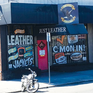 A storefront with a black awning that reads JUST LEATHER. The window displays signs advertising leather jackets, vests, boots, chaps, and alterations. A motorcycle is parked in front of the shop.