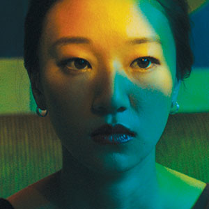 A woman with straight dark hair looks ahead with a neutral expression. Colored lights in yellow, green, and blue cast dramatic shadows across her face. She wears small hoop earrings and a dark top.