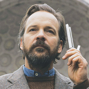 A man with a beard and mustache holds two metal rods near his face. He wears a brown jacket over a blue shirt and is standing outdoors in front of a patterned background.