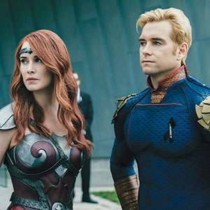 Two people in superhero costumes stand side by side. The woman has long red hair and wears silver and maroon armor, while the man has short blond hair and wears a blue and gold suit. They look serious and focused.