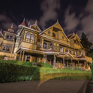 A large, ornate Victorian mansion with multiple peaked roofs, turrets, and decorative trim, illuminated at night against a cloudy sky, with manicured hedges in the foreground.