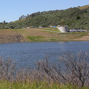 A reservoir with calm blue water in the foreground, a grassy embankment and dry shrubs, with a concrete spillway structure and vehicles visible on the road near green hills in the background.