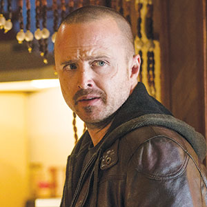 A man with closely shaved hair and a short beard wears a brown leather jacket and hoodie, looking off to the side with a serious expression. Beaded curtains hang in the background.