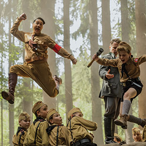 A group of boys in brown uniforms stack together in a forest. One man in a similar uniform leaps into the air beside a jumping boy, both energetic and animated, while another man stands behind them watching.