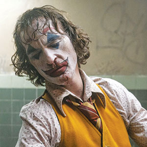 A person with messy hair and dramatic clown makeup, including a red painted mouth and blue shapes above and below the eyes, wears a yellow vest, patterned shirt, and striped tie, standing in a dimly lit tiled room.