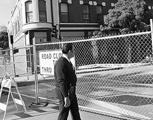 A man in a suit walks past a chain-link fence blocking the street, with a Road Closed to Thru Traffic sign visible behind him. A building and trees can be seen in the background.