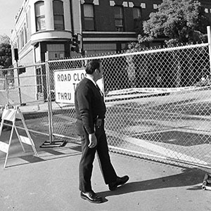 A man in a suit walks past a chain-link fence blocking the street, with a Road Closed to Thru Traffic sign visible behind him. A building and trees can be seen in the background.