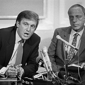 Two men in suits sit at a table with several microphones in front of them. One man is speaking and gesturing with his hands, while the other looks on attentively. The image is in black and white.