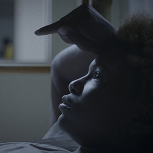 A person lies in bed, gazing upward with their hand resting on their forehead, illuminated by soft, dim light from a nearby window.