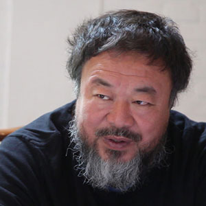 A middle-aged man with a full beard and mustache, dark hair, and wearing a dark shirt, sits indoors and looks slightly to the side, speaking or listening attentively.
