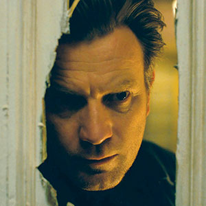 A man with intense expression peers through a jagged hole in a white wooden door, his face partially lit by a warm light from behind.