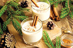 Two glasses of eggnog topped with cinnamon sticks sit on a rustic wooden surface, surrounded by evergreen branches, pine cones, cinnamon sticks, and a small dish of ground cinnamon.