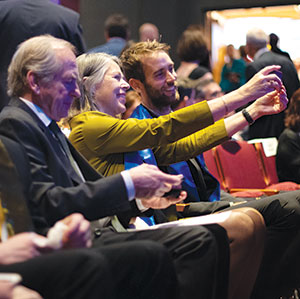 Three people sit in an audience; a woman in a yellow top and a man beside her are smiling and taking a selfie together, while an older man in a suit sits to their left, looking ahead. Other attendees are visible in the background.