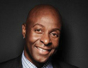 A smiling man in a dark suit, blue checkered shirt, and blue tie poses against a dark background. He has a shaved head and is wearing a small earring in his left ear.