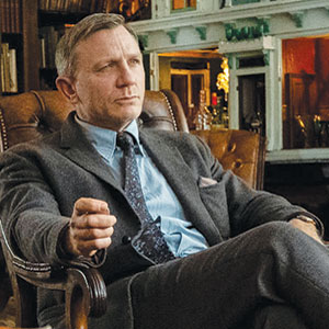 A man in a gray suit sits in a leather armchair, looking thoughtful. He wears a blue shirt and patterned tie, with one hand resting on the chairs arm. The background features shelves and warm lighting.