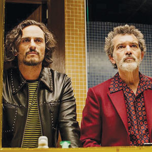 Two men stand side by side, looking into a mirror. One wears a black leather jacket over a striped shirt, the other wears a red blazer and patterned shirt. The background features yellow and blue tiled walls.