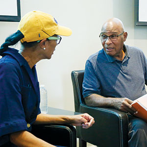 Two people sit and talk indoors. One, wearing a yellow cap and glasses, faces a man in a blue shirt holding an open book. They appear engaged in a conversation. A water bottle sits on a table behind them.