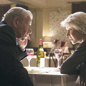 An older man and woman sit across from each other at a restaurant table, engaged in a serious conversation. Wine glasses and a bottle of wine are on the table, with other diners blurred in the background.