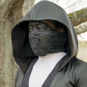 A person wearing a black hooded cloak and a matching black face mask stands outdoors. The background shows tree branches with no leaves. The persons eyes are visible, and they are dressed in dark, concealing clothing.