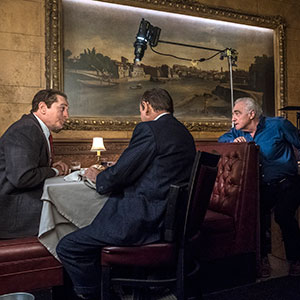 Three men sit at a restaurant table in a booth, engaged in conversation. A large painting is on the wall behind them, and a boom microphone is visible above, indicating a film set.