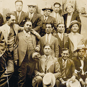 A sepia-toned vintage photo of a group of men in suits and hats, posing closely together and looking at the camera, with some standing and others seated.