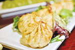 A close-up of three golden-brown dumplings arranged on a white rectangular plate, garnished with fresh greens. Another dish is blurred in the background.