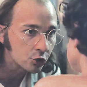 A man with long hair, glasses, and sideburns leans forward, speaking intensely to someone whose back is facing the camera.