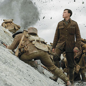A group of soldiers in brown uniforms climb a dirt slope during battle; one stands upright looking ahead as an explosion sends debris into the air behind them.