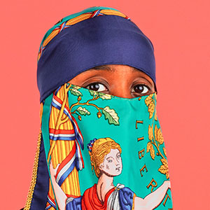 A person wearing a colorful scarf covering their head and lower face, with only their eyes visible. The scarf features an illustrated woman, leaves, and the word LIBERTY on a bright, solid pink background.