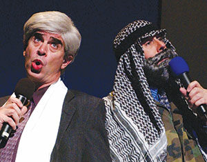 Two men on stage, one dressed as a Jewish man with a white tallit and gray wig, the other as an Arab man in a keffiyeh and camouflage, both holding microphones and singing or speaking animatedly.