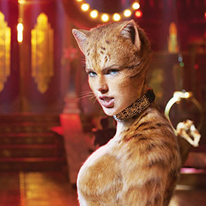 A person dressed in a detailed orange cat costume with fur, whiskers, and cat ears stands in a theatrical, colorful, and dramatic stage setting with glowing lights in the background.