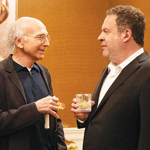Two men in suits stand facing each other indoors, engaged in conversation. One holds a bottle, the other holds a glass with a drink. The background is warmly lit with brown and gold tones.