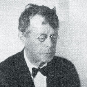 Black and white photo of an older man wearing round glasses, a suit, and a bow tie. He has short, slightly messy hair and appears to be looking down with a neutral expression.